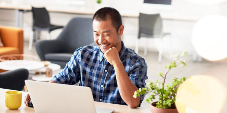 Young Asian designer smiling confidently while sitting alone at a desk in a modern office working on a laptop; Shutterstock ID 627947441; Purchase Order: 20076173; Job: SL adviser images