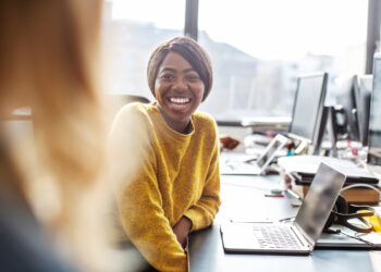 Two young business women working together in office, Female professionals sitting at the desk talking with each other and smiling.