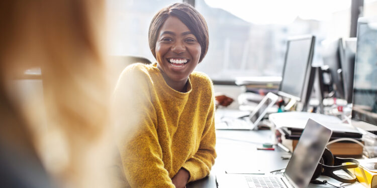Two young business women working together in office, Female professionals sitting at the desk talking with each other and smiling.
